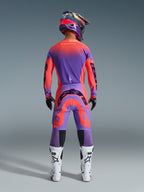 Pantalones Alpinestars Supertech Scenz 2026, pantalones de motocross en coral intenso y púrpura, vista trasera de un piloto de motocross con un conjunto completo de equipamiento de carreras, diseño con degradado vibrante y logotipos traseros llamativos, combinados con jersey a juego, casco y botas blancas