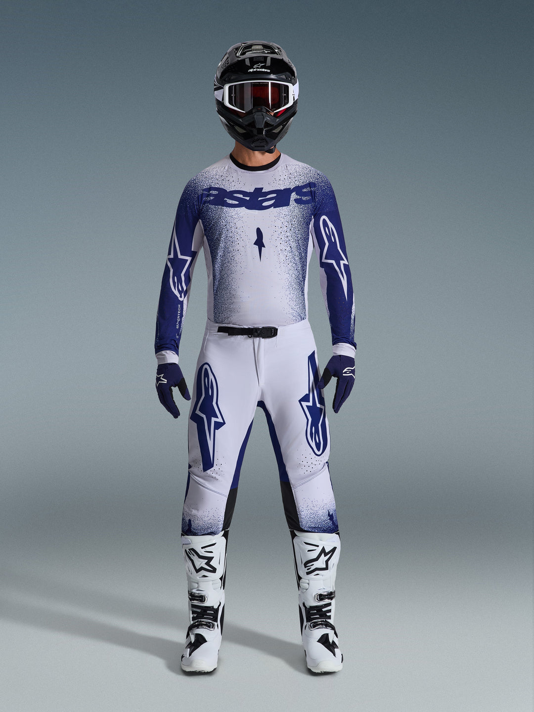 Piloto de motocross con el conjunto de equipamiento Alpinestars 2026 Supertech Scenz, jersey blanco y azul oscuro con pantalones a juego, presenta logotipos grandes de Astars y un diseño gráfico moteado, combinado con un casco negro, botas protectoras blancas y negras, y guantes azules coordinados, equipo de competición profesional completo.