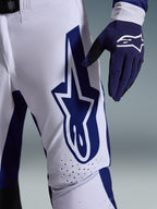 Alpinestars 2026 Supertech Scenz Pants, Motocross Pants, Light Gray and Dark Blue, close-up view of the thigh showing a large dark blue Alpinestars logo, perforated ventilation zones, and stretch rip-stop fabric, worn with a matching dark blue motocross glove