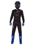 Alpinestars 2025 Racer Graphite Pants, pantalones de motocross, negro y gris, usados por un modelo con un jersey negro a juego, botas azules y moradas, y gafas azules, conjunto completo de carreras off-road con logotipos llamativos de Alpinestars