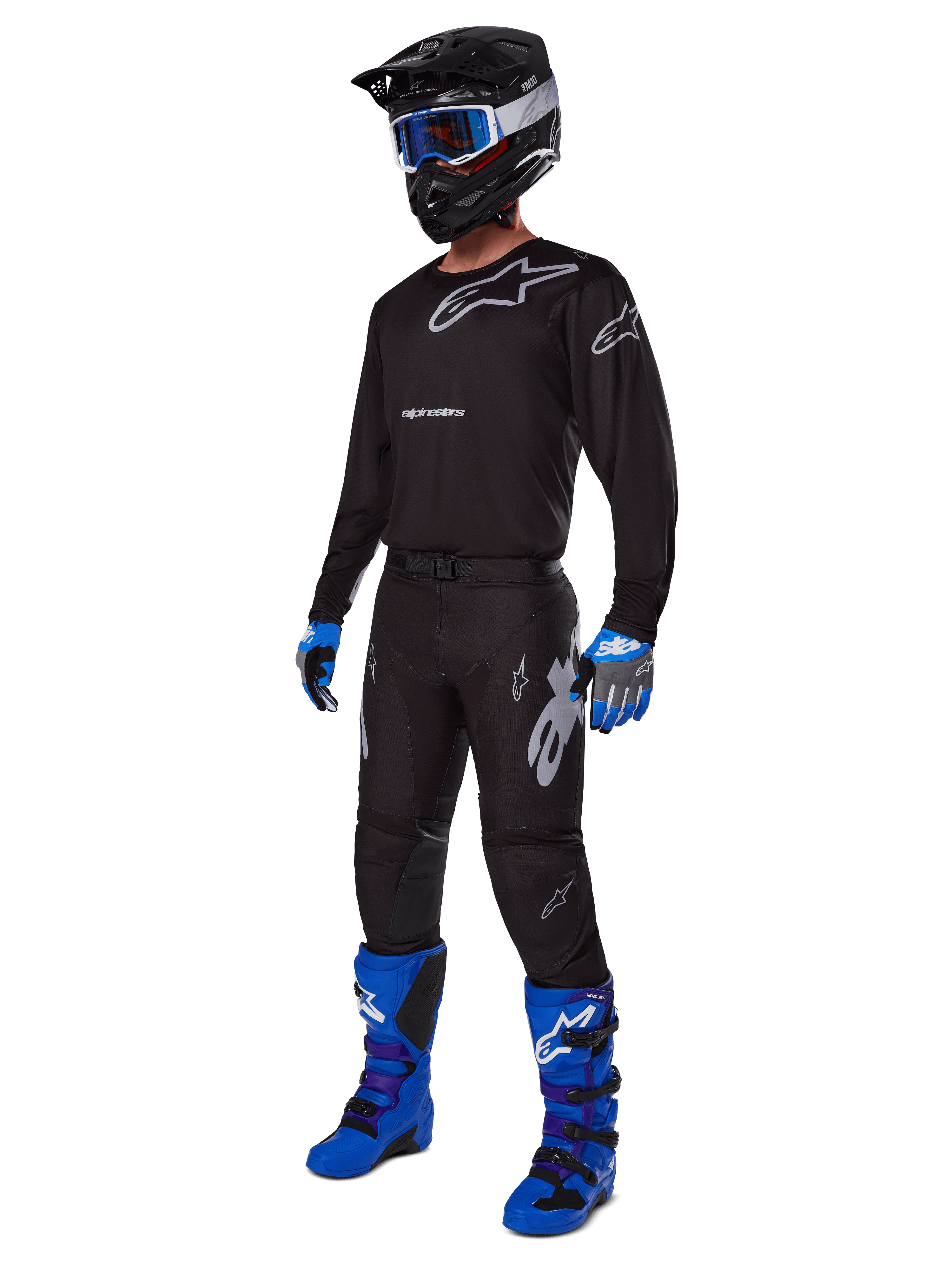 Piloto de motocross con equipamiento de carreras completo de Alpinestars, pantalones Racer Graphite 2025 en negro y gris, jersey negro a juego con logotipos grises, casco negro con gafas azules, botas de color azul brillante y púrpura, y guantes azules y grises, conjunto coordinado para carreras off-road