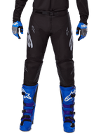 Alpinestars 2025 Racer Graphite Pants, pantalones de motocross, negros y grises, usados con guantes azules y grises y botas azules, con marca Astars plateada en los muslos y rodillas reforzadas, diseño deportivo para rendimiento off-road
