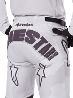 Vista trasera de los pantalones Alpinestars Racer Hollow 2025, pantalones de motocross, blanco y camuflaje claro, con el logotipo negro de Alpinestars en el asiento, usados con un jersey a juego y guantes negros