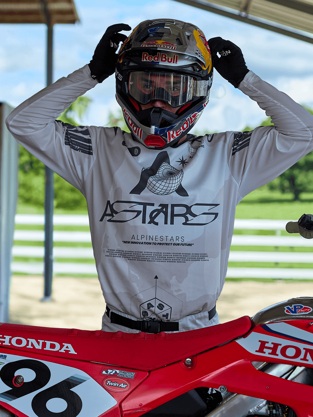 Piloto de motocross vistiendo equipamiento Alpinestars Racer Hollow 2025, jersey de camuflaje claro con el logotipo ASTARS destacado, ajustando un casco Red Bull con gafas transparentes, junto a una moto de motocross Honda roja en un entorno todoterreno al aire libre.