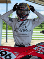 Piloto de motocross vistiendo equipamiento Alpinestars Racer Hollow 2025, jersey de camuflaje claro con el logotipo ASTARS destacado, ajustando un casco Red Bull con gafas transparentes, junto a una moto de motocross Honda roja en un entorno todoterreno al aire libre.