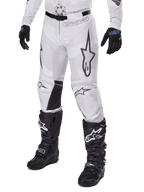 Alpinestars 2025 Racer Hollow Pants, pantalones de motocross, camuflaje blanco y gris, con grandes logotipos de estrellas contorneados en negro y paneles interiores de cuero negro en las rodillas, usados con botas y guantes de motocross negros, diseño duradero para carreras off-road.