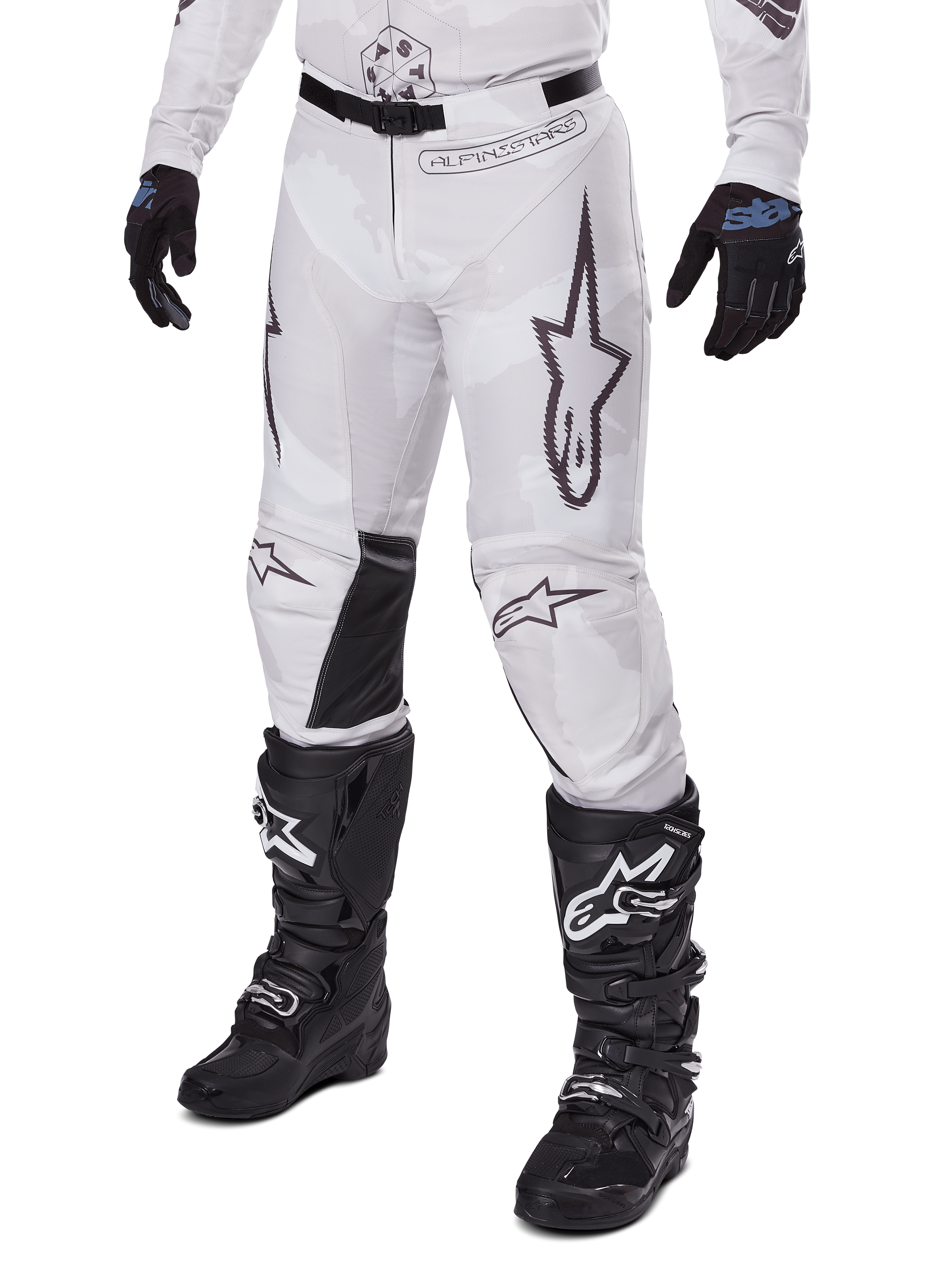 Alpinestars 2025 Racer Hollow Pants, pantalones de motocross, camuflaje blanco y gris, con grandes logotipos de estrellas contorneados en negro y paneles interiores de cuero negro en las rodillas, usados con botas y guantes de motocross negros, diseño duradero para carreras off-road.