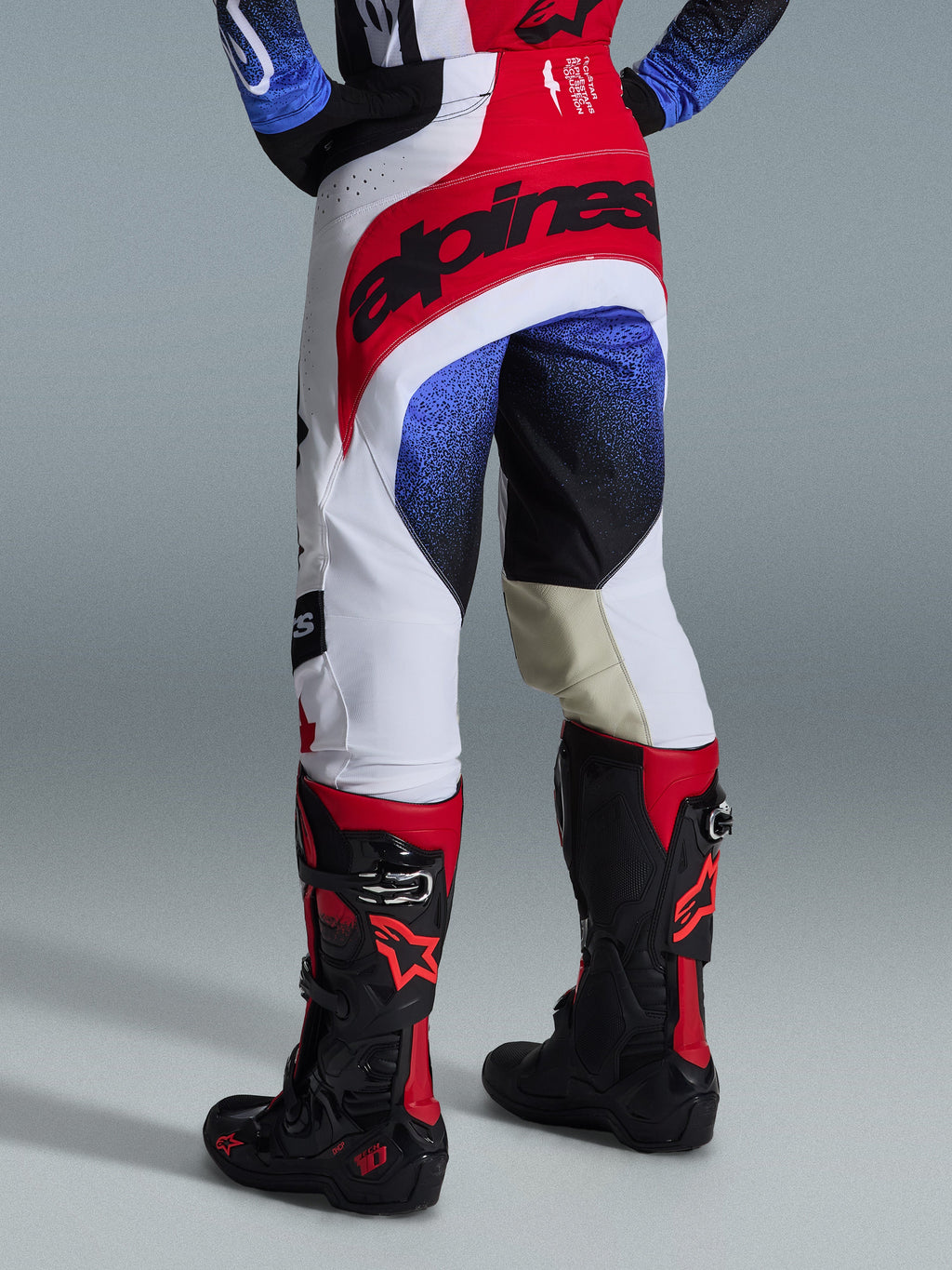 Pantalones Alpinestars Techstar Knif 2026, pantalones de motocross, blanco, rojo y azul, vista trasera que muestra el asiento rojo con marca negra, patrones moteados azules y negros en las piernas, usados con botas Tech 10 negras y rojas, equipamiento de competición off-road de élite