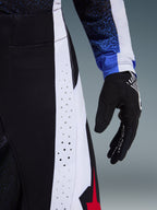 Detalle en primer plano de los pantalones Alpinestars Techstar Knif 2026 en color negro, blanco y rojo, que muestra las perforaciones cortadas por láser en un panel lateral blanco para la ventilación, presentados con un guante de motocross negro y la manga de un jersey azul estampado, destacando su construcción de alto rendimiento para competición.
