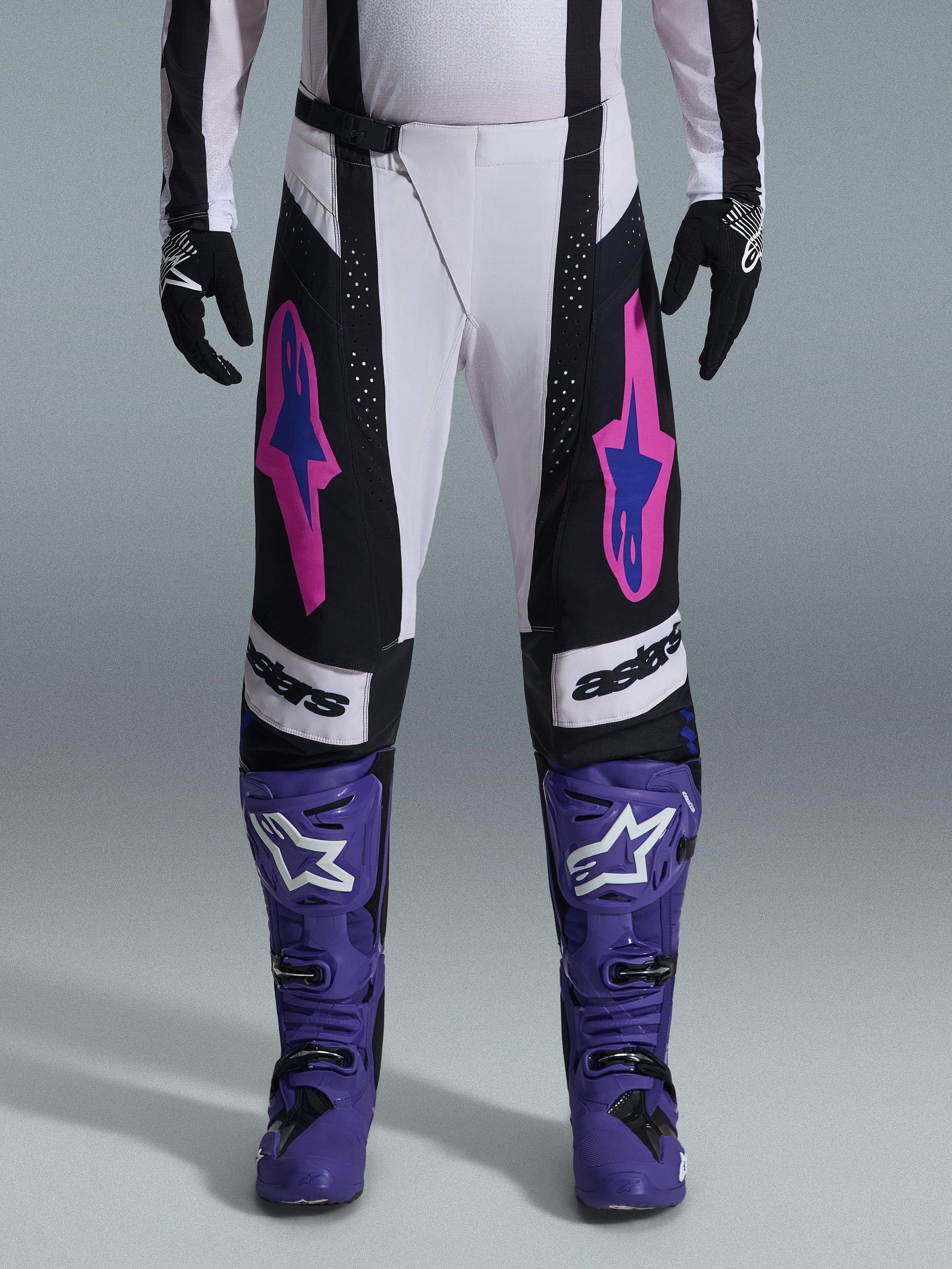 Alpinestars 2026 Techstar Knif Pants, pantalones de motocross, en negro, blanco y morado con detalles en rosa y azul, usados con botas de carreras moradas y guantes negros, con un corte de competición ergonómico y logotipos llamativos para rendimiento off-road