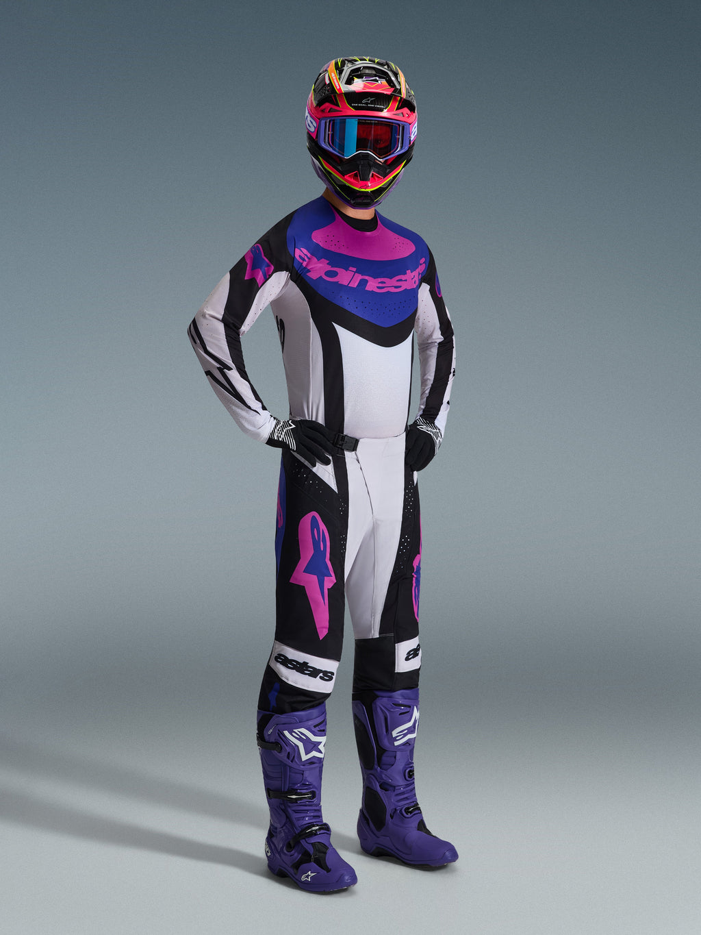 Motocross rider wearing the Alpinestars 2026 Techstar Knif Gear Set, Black Light Gray and Purple, white jersey with blue and pink accents, matching pants with purple star logos, colorful pink and yellow helmet, purple motocross boots, and black gloves, professional gear set for off-road racing.