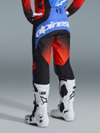 Pantalones Alpinestars Techstar Knif 2026, pantalones de motocross en naranja, negro y azul UCLA, vista trasera que muestra el canesú azul con el logotipo blanco de Alpinestars, patrón de degradado naranja en las piernas, usados con camiseta a juego y botas blancas para carreras de motocross