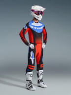 Piloto de motocross con los pantalones Techstar Knif 2026 en naranja, negro y azul UCLA, combinados con un jersey de carreras a juego, casco blanco con gafas rojas y azules, y botas de motocross blancas; conjunto completo de equipamiento profesional para motocross.