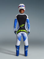 Piloto de motocross de espaldas con equipamiento de carreras completo, pantalones Techstar Knif 2026 en UCLA azul, negro y amarillo flúor, jersey de manga larga a juego con paneles negros y blancos, botas azules, casco blanco y el logotipo de Alpinestars en amarillo flúor en el asiento.