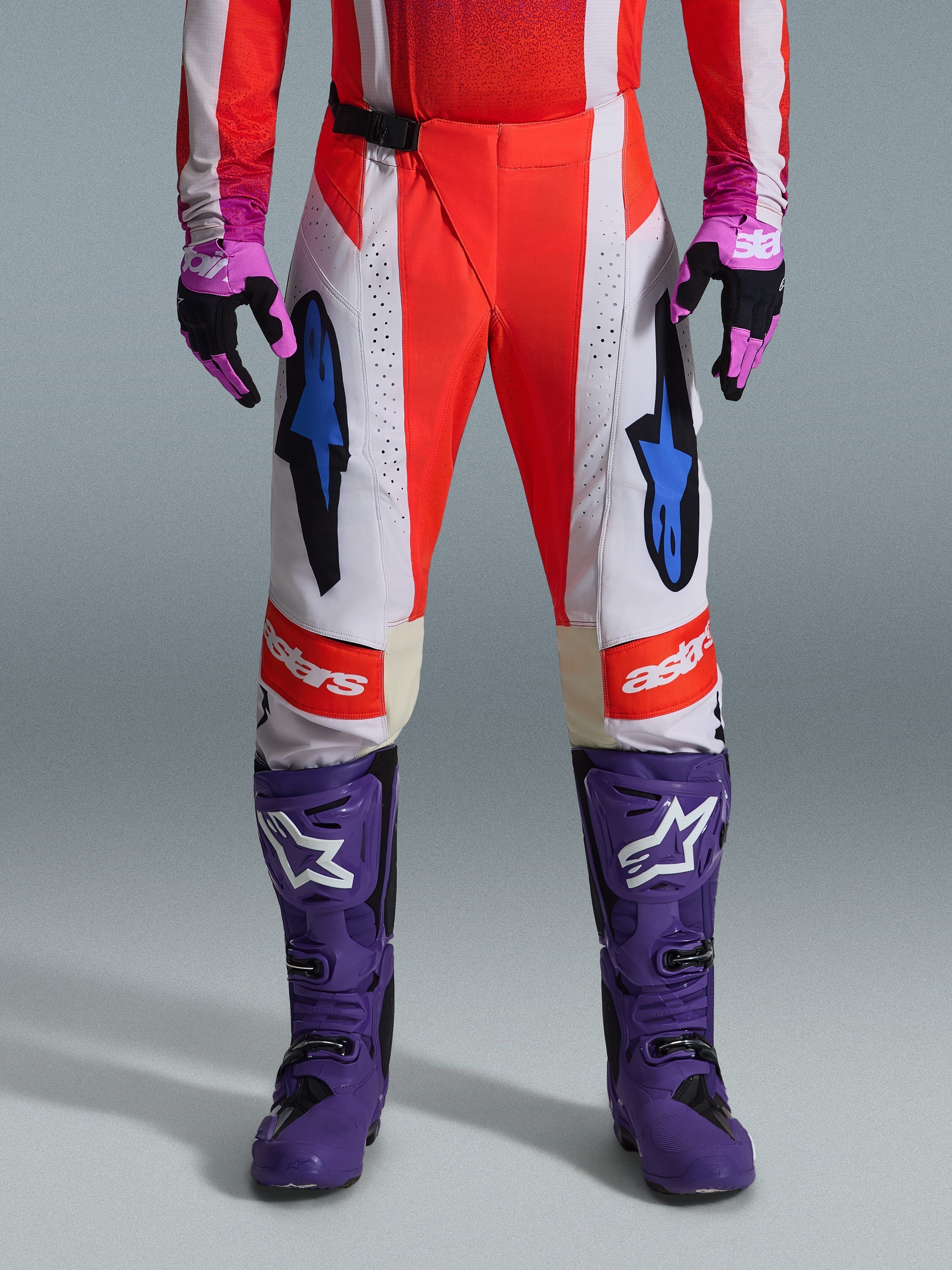 Alpinestars Techstar Knif Pants 2026, pantalones de motocross en naranja flúor y gris claro con detalles en negro, mostrados con botas y guantes de motocross morados, con logotipos de estrellas azules y construcción de paneles múltiples para rendimiento todoterreno