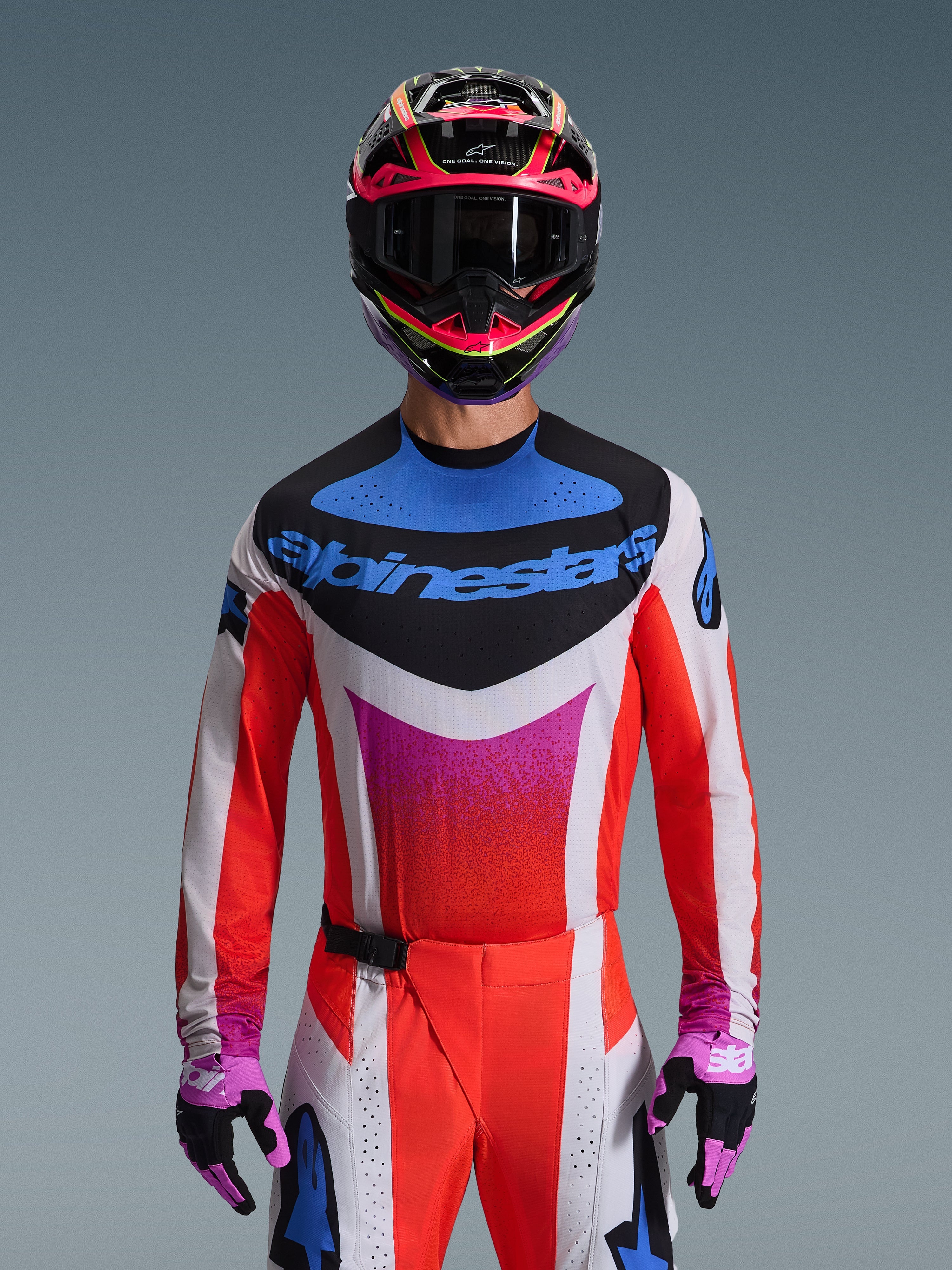 Piloto de motocross con el jersey Alpinestars 2026 Techstar Knif en color gris claro, naranja flúor y negro, con un llamativo logotipo azul y vibrantes mangas naranjas, combinado con un casco rosa y negro y guantes morados, ajuste atlético de carrera para competición todoterreno de alto rendimiento.