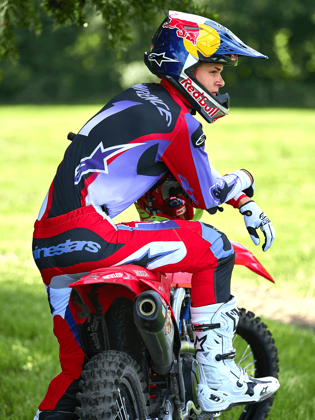 Piloto de motocross sentado en una moto de cross roja, vistiendo pantalones Alpinestars 2025 Fluid Wurx en púrpura y rojo brillante, jersey a juego, botas Tech 10 en blanco y negro, guantes blancos y casco de Red Bull, conjunto de carreras off-road completo en un entorno al aire libre