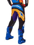 Alpinestars 2025 Fluid Wurx Pants, pantalones de motocross, multicolor con naranja, morado y azul, vista trasera que muestra el logotipo blanco de Alpinestars en la parte posterior, combinados con botas de motocross azules, ajuste atlético para carreras todoterreno