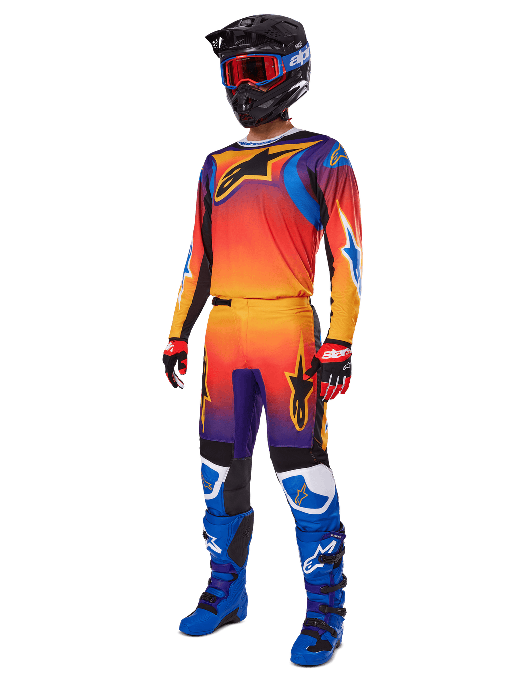 Piloto de motocross con equipamiento completo de carreras, pantalones Alpinestars 2025 Fluid Wurx y jersey a juego en un degradado multicolor naranja, rojo y morado, casco negro con gafas rojas, botas azules y negras, y guantes rojos y negros, conjunto coordinado para carreras todoterreno