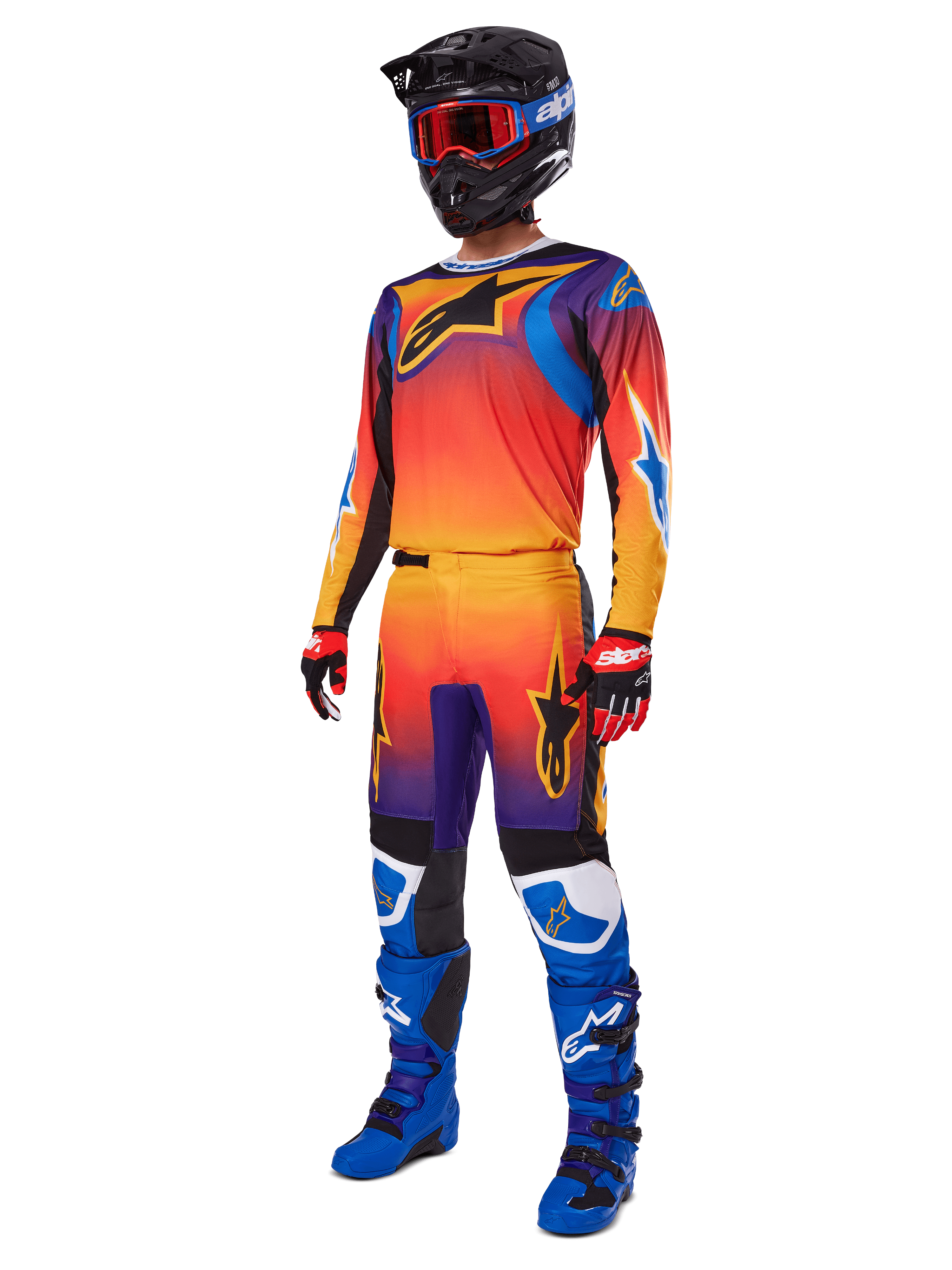 Piloto de motocross con equipamiento completo de carreras, pantalones Alpinestars 2025 Fluid Wurx y jersey a juego en un degradado multicolor naranja, rojo y morado, casco negro con gafas rojas, botas azules y negras, y guantes rojos y negros, conjunto coordinado para carreras todoterreno