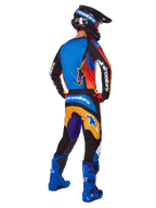 Piloto de motocross visto desde atrás con pantalones Alpinestars Fluid Wurx 2025, diseño multicolor con detalles en azul, naranja, amarillo y púrpura, combinados con un jersey a juego, botas azul brillante y casco negro, equipamiento completo de competición off-road