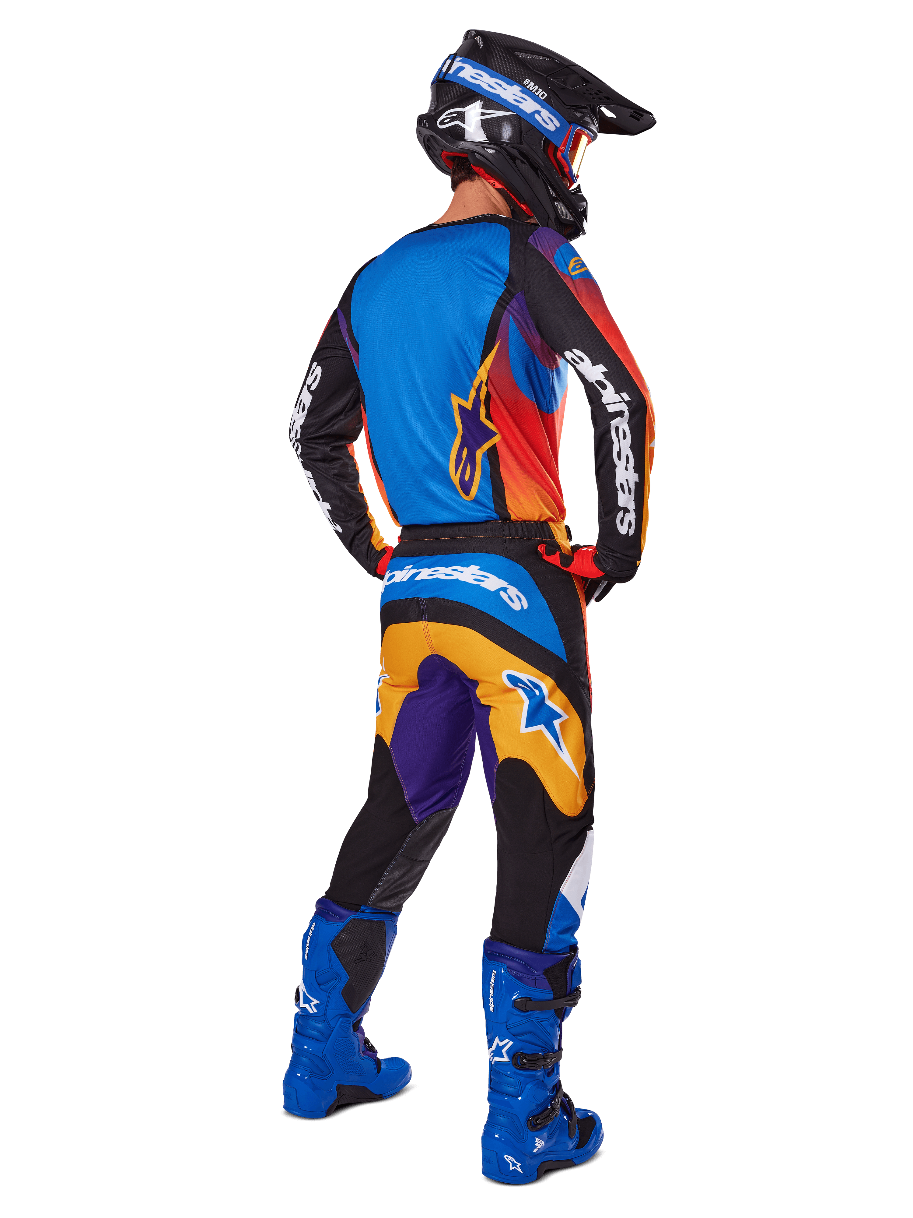 Piloto de motocross visto desde atrás con pantalones Alpinestars Fluid Wurx 2025, diseño multicolor con detalles en azul, naranja, amarillo y púrpura, combinados con un jersey a juego, botas azul brillante y casco negro, equipamiento completo de competición off-road