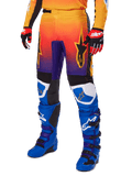 Alpinestars 2025 Fluid Wurx Pants, pantalones de motocross, degradado multicolor en naranja, rojo y púrpura con paneles negros y azules, usados con botas azules y guantes rojos, con gráficos del logo de estrella y detalles blancos en las rodillas, equipamiento de alto rendimiento para carreras todoterreno