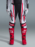 Alpinestars 2026 Techstar Nomur Pants, pantalones de motocross, negro, blanco y rojo, combinados con botas rojas a juego y guantes blancos, con logotipos rojos llamativos en los muslos y marca 'astars' en las rodillas, diseño de competición de paneles múltiples para rendimiento todoterreno