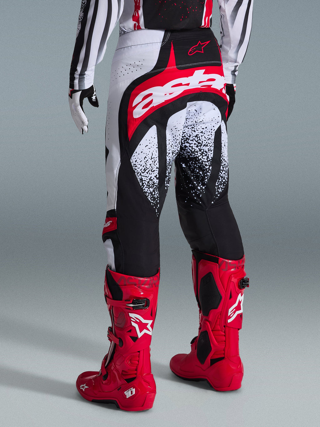 Alpinestars Techstar Nomur Pants 2026, pantalones de motocross, negros, blancos y rojos, vista posterior en un piloto con botas rojas a juego, con un llamativo logotipo 'astars' en rojo y blanco en el asiento y patrones gráficos moteados, equipamiento profesional de carreras todoterreno