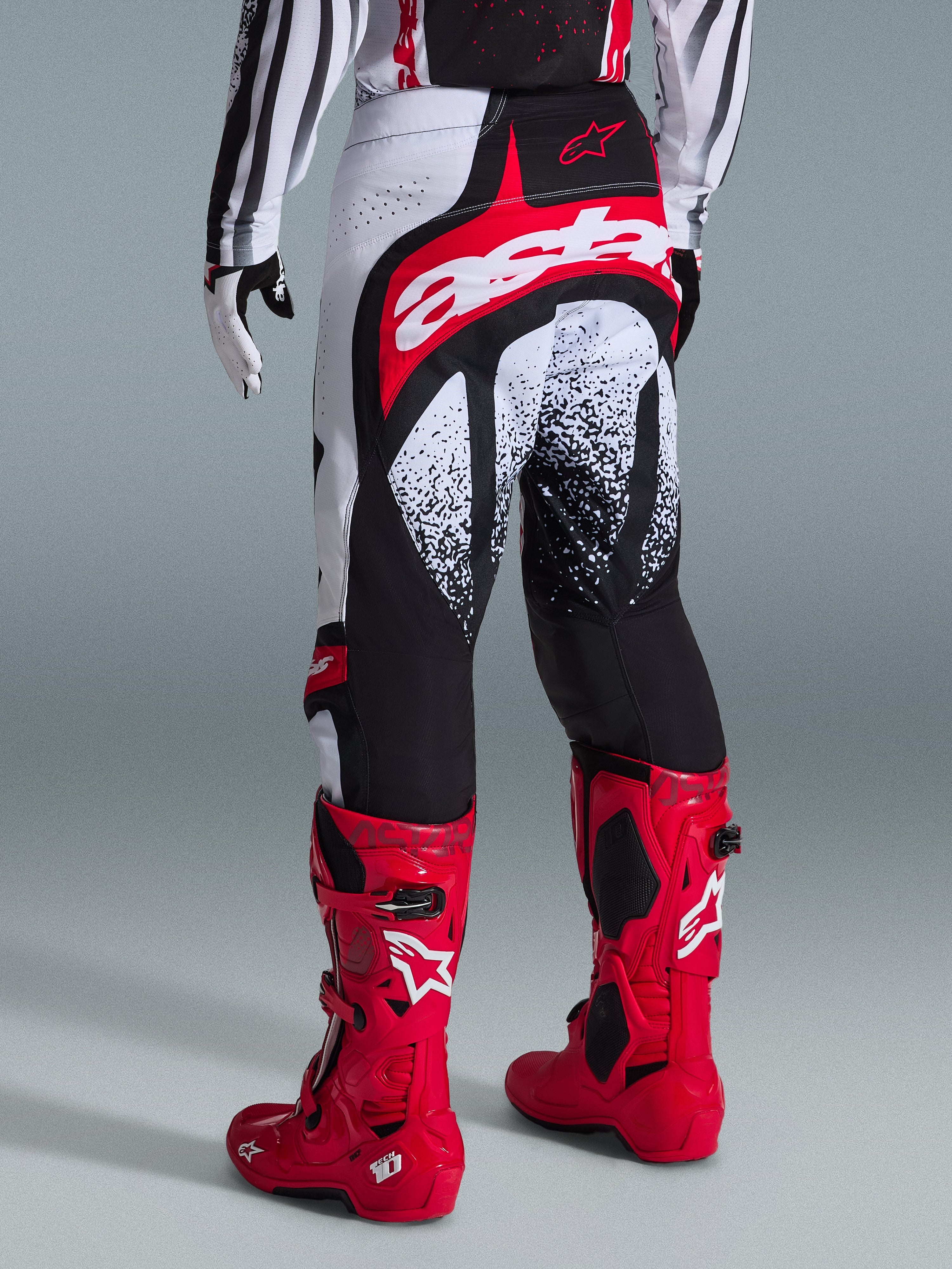 Alpinestars Techstar Nomur Pants 2026, pantalones de motocross, negros, blancos y rojos, vista posterior en un piloto con botas rojas a juego, con un llamativo logotipo 'astars' en rojo y blanco en el asiento y patrones gráficos moteados, equipamiento profesional de carreras todoterreno