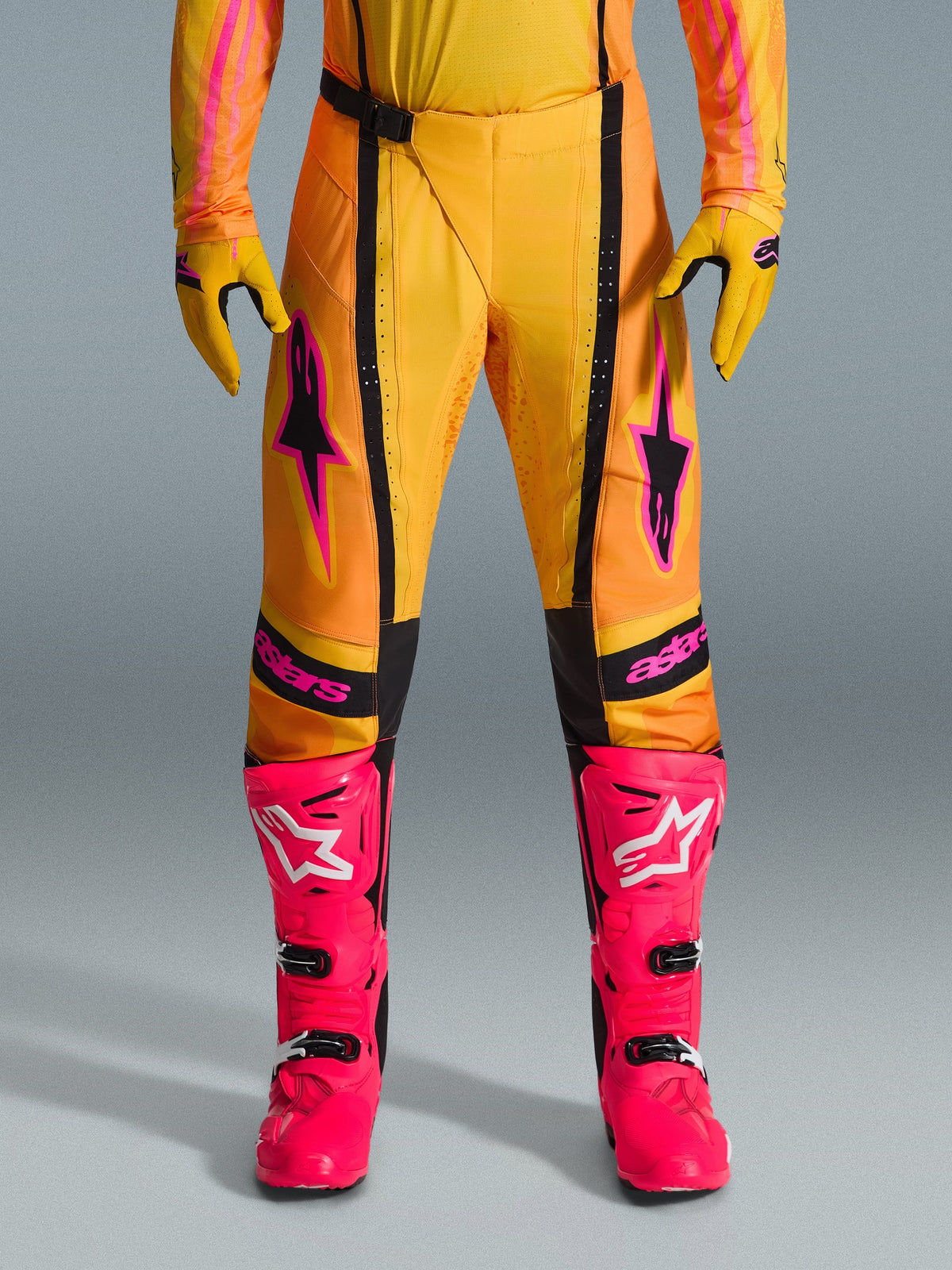 Alpinestars Techstar Nomur 2026, pantalones de motocross, naranja, amarillo y rosa, combinados con guantes amarillos a juego y botas de competición rosa brillante, con paneles negros y marca Alpinestars en rosa, equipamiento técnico de carreras todoterreno
