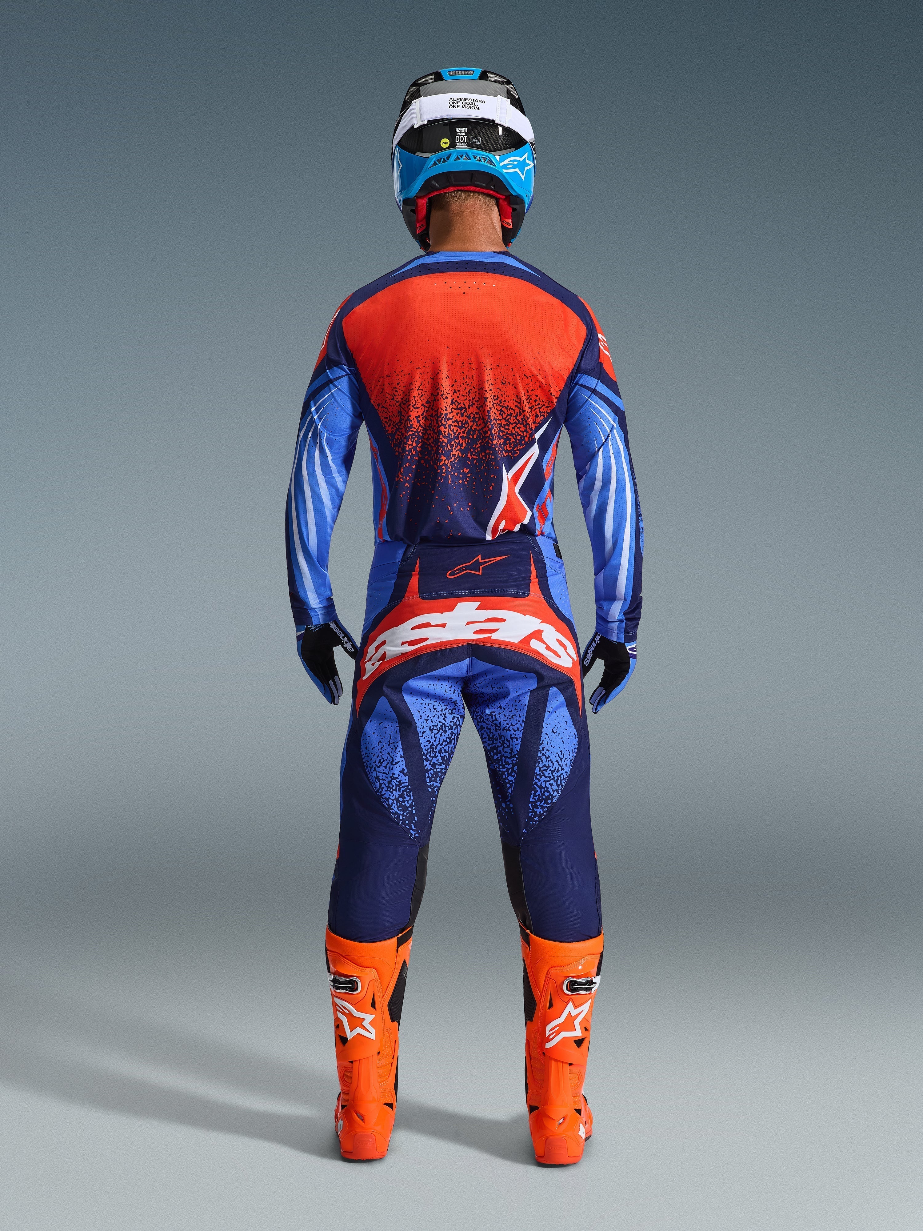 Motocross rider seen from the back wearing Alpinestars 2026 Techstar Nomur Pants in Dark Navy, Orange, and UCLA Blue, styled with a matching jersey and bright orange boots, professional off-road racing gear set.