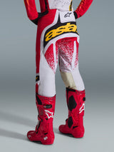 Vista trasera de un piloto de motocross con pantalones Alpinestars 2026 Techstar Nomur en rojo y blanco, con un llamativo logo amarillo en el asiento, combinados con botas de motocross Tech 10 en color rojo brillante, toma de estudio sobre fondo gris.