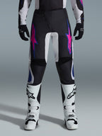 Pantalones Alpinestars 2026 Fluid Grid, pantalones de motocross, negros con diseños de estrellas en púrpura y azul, combinados con botas de motocross blancas y guantes negros, con paneles reforzados y un ajuste deportivo off-road, fondo gris