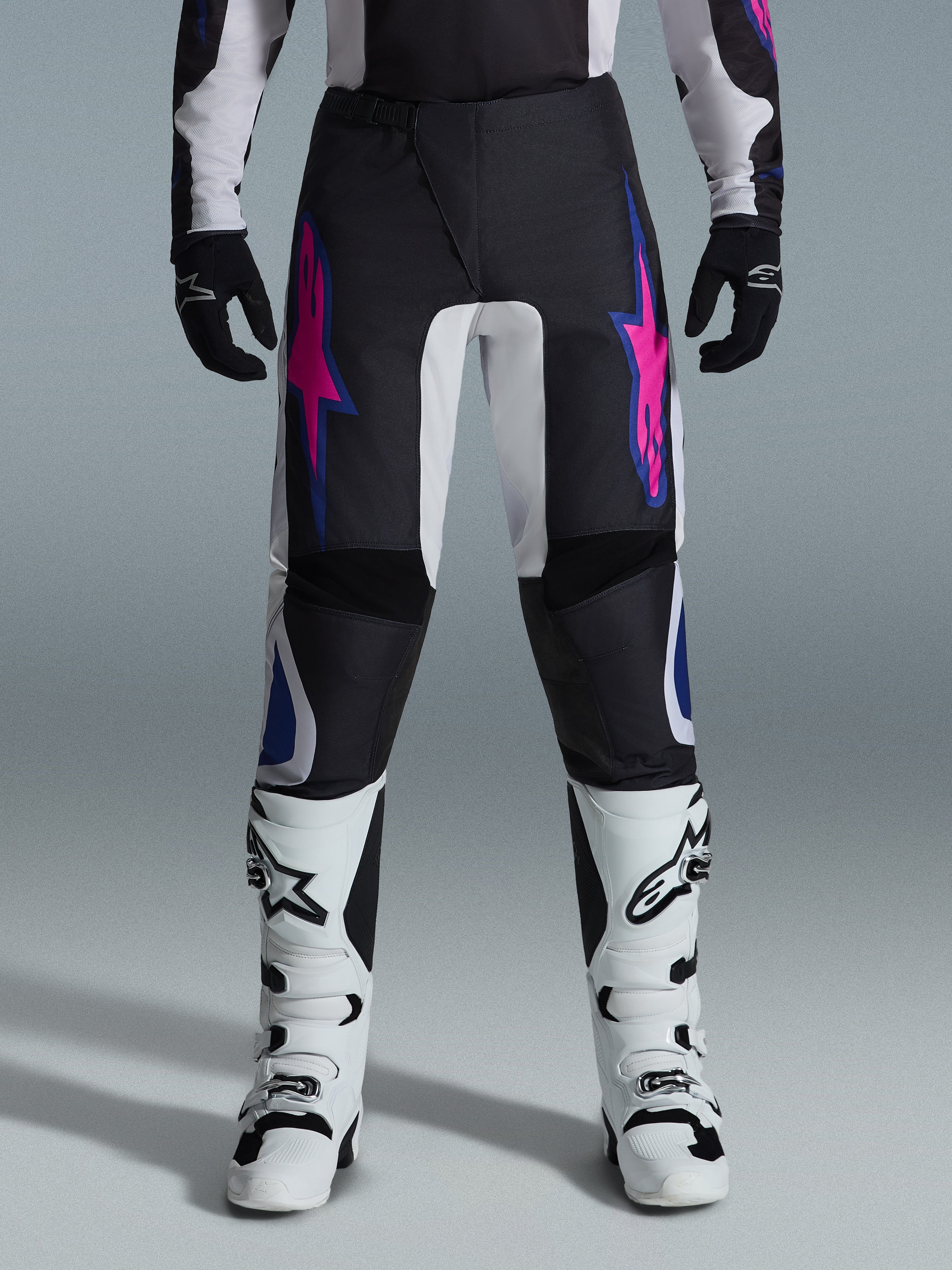 Pantalones Alpinestars 2026 Fluid Grid, pantalones de motocross, negros con diseños de estrellas en púrpura y azul, combinados con botas de motocross blancas y guantes negros, con paneles reforzados y un ajuste deportivo off-road, fondo gris