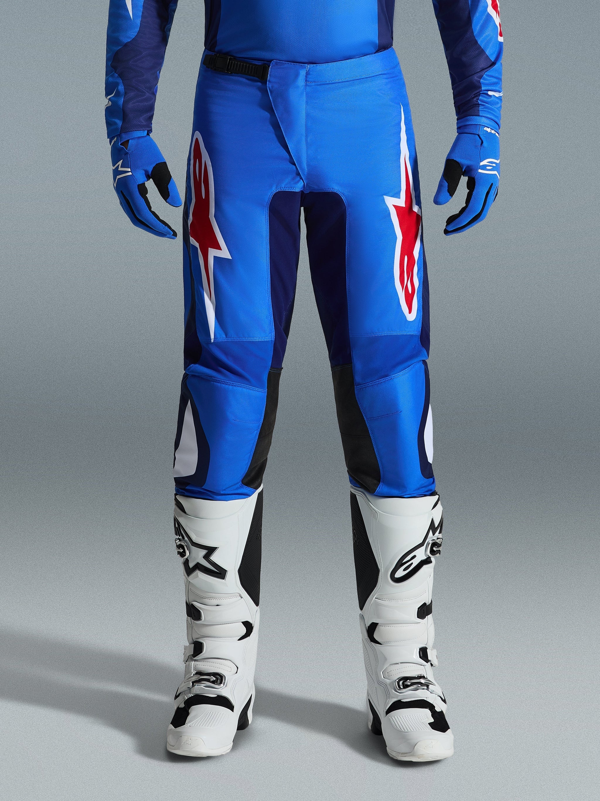 Alpinestars 2026 Fluid Grid Pants, pantalones de motocross, azul y rojo, con llamativos logotipos de estrellas rojos y blancos en los muslos, combinados con guantes azules a juego y botas blancas de off-road, ajuste deportivo con paneles resistentes para competición.