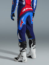 Alpinestars 2026 Fluid Grid Pants, pantalones de motocross, azul y rojo, vista trasera que muestra la marca Alpinestars destacada y el diseño ergonómico, usados con botas Tech 7 blancas y negras y jersey a juego, equipamiento de alto rendimiento para conducción off-road