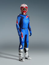 Piloto de motocross con equipamiento de carreras completo, jersey azul y rojo con patrón de cuadrícula y logo de Alpinestars, pantalones Fluid Grid 2026 en azul y rojo con paneles internos negros en las rodillas, casco rojo y blanco con gafas, botas blancas y negras, guantes azules, conjunto coordinado para carreras off-road