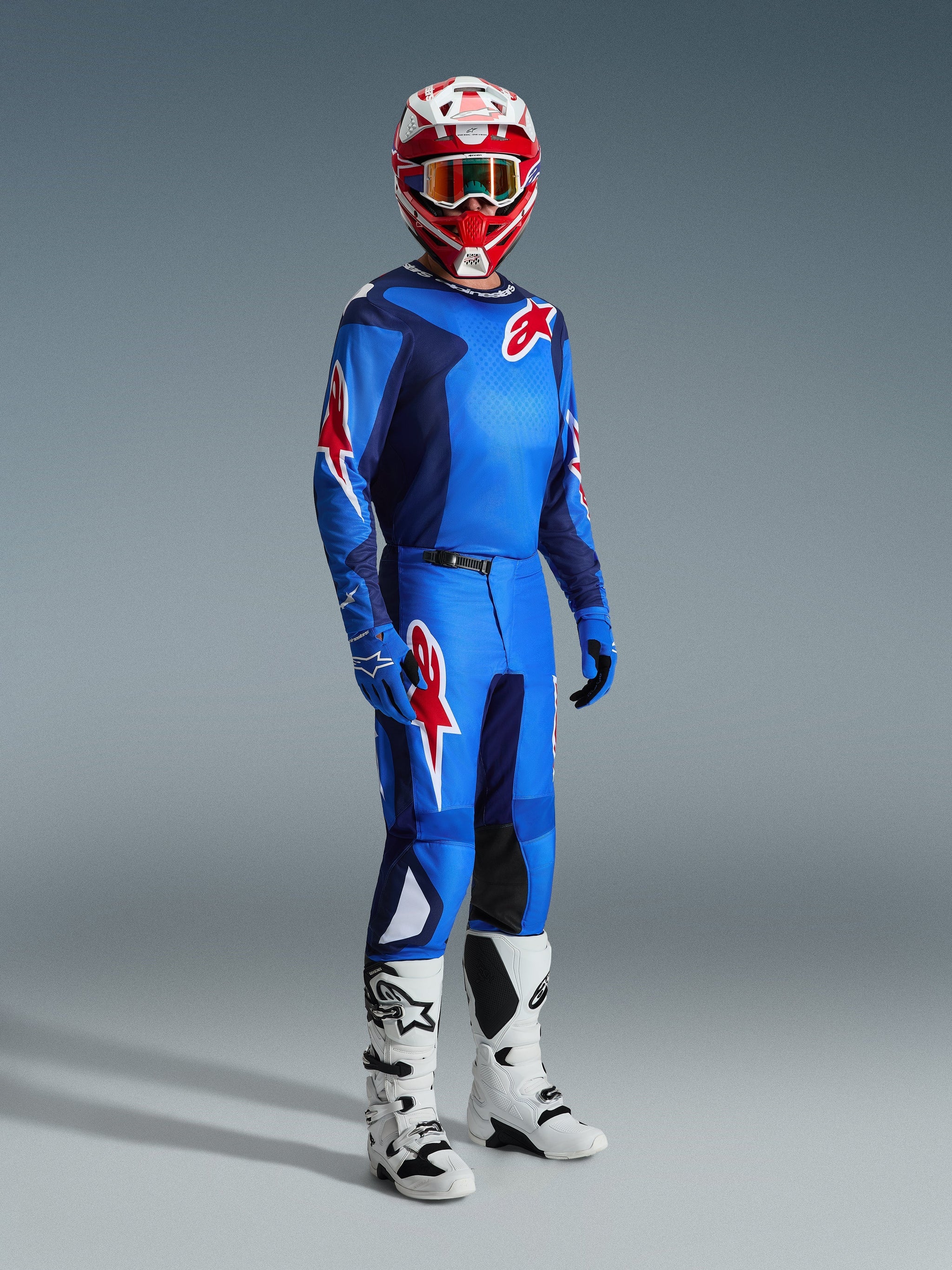 Piloto de motocross con equipamiento de carreras completo, jersey azul y rojo con patrón de cuadrícula y logo de Alpinestars, pantalones Fluid Grid 2026 en azul y rojo con paneles internos negros en las rodillas, casco rojo y blanco con gafas, botas blancas y negras, guantes azules, conjunto coordinado para carreras off-road