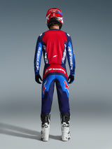Piloto de motocross visto desde atrás con equipamiento completo de competición, pantalones Fluid Grid 2026 en azul y rojo, jersey a juego con la marca Alpinestars, casco rojo y azul, botas blancas y negras, conjunto coordinado para carreras off-road