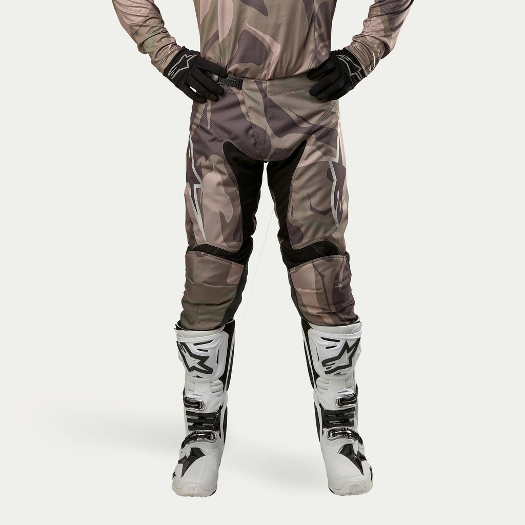 Alpinestars Racer Tactical Pants 2024, pantalones de motocross, verde camuflaje militar marrón, con grandes logotipos Alpinestars blancos en los muslos y paneles interiores de las piernas negros, combinados con botas de motocross blancas y negras, guantes negros y jersey de camuflaje a juego, vista frontal.