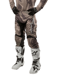 Alpinestars 2024 Racer Tactical Pants, pantalones de motocross en camuflaje verde militar y marrón, usados con jersey de camuflaje a juego, guantes negros y botas blancas con logos de estrella, con paneles elásticos negros y diseño ergonómico, equipo para todoterreno sobre fondo blanco.
