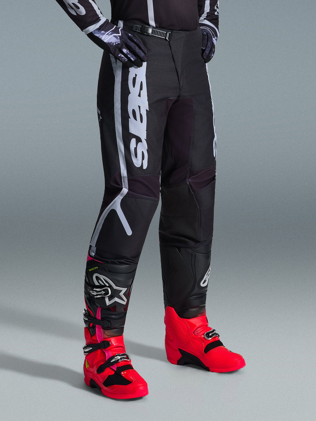 Pantalones Alpinestars Fluid Apex 2026, pantalones de motocross, negros y grises, usados con botas de color rosa vibrante y rojo y jersey a juego en blanco y negro, con el logotipo vertical de Alpinestars en el muslo, paneles reforzados y cierre de trinquete en la cintura para un óptimo rendimiento off-road.