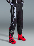 Pantalones Alpinestars Fluid Apex 2026, pantalones de motocross, negros y grises, usados con botas de color rosa vibrante y rojo y jersey a juego en blanco y negro, con el logotipo vertical de Alpinestars en el muslo, paneles reforzados y cierre de trinquete en la cintura para un óptimo rendimiento off-road.