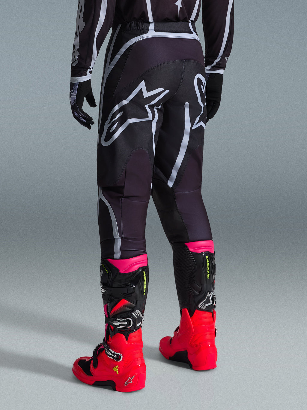 Pantalones Alpinestars Fluid Apex 2026, pantalones de motocross, negro y gris, vista trasera con grandes logotipos blancos de Alpinestars en el asiento, usados con botas en rojo brillante y rosa, construcción duradera de polifabric 600D para rendimiento off-road