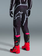 Pantalones Alpinestars Fluid Apex 2026, pantalones de motocross, negro y gris, vista trasera con grandes logotipos blancos de Alpinestars en el asiento, usados con botas en rojo brillante y rosa, construcción duradera de polifabric 600D para rendimiento off-road