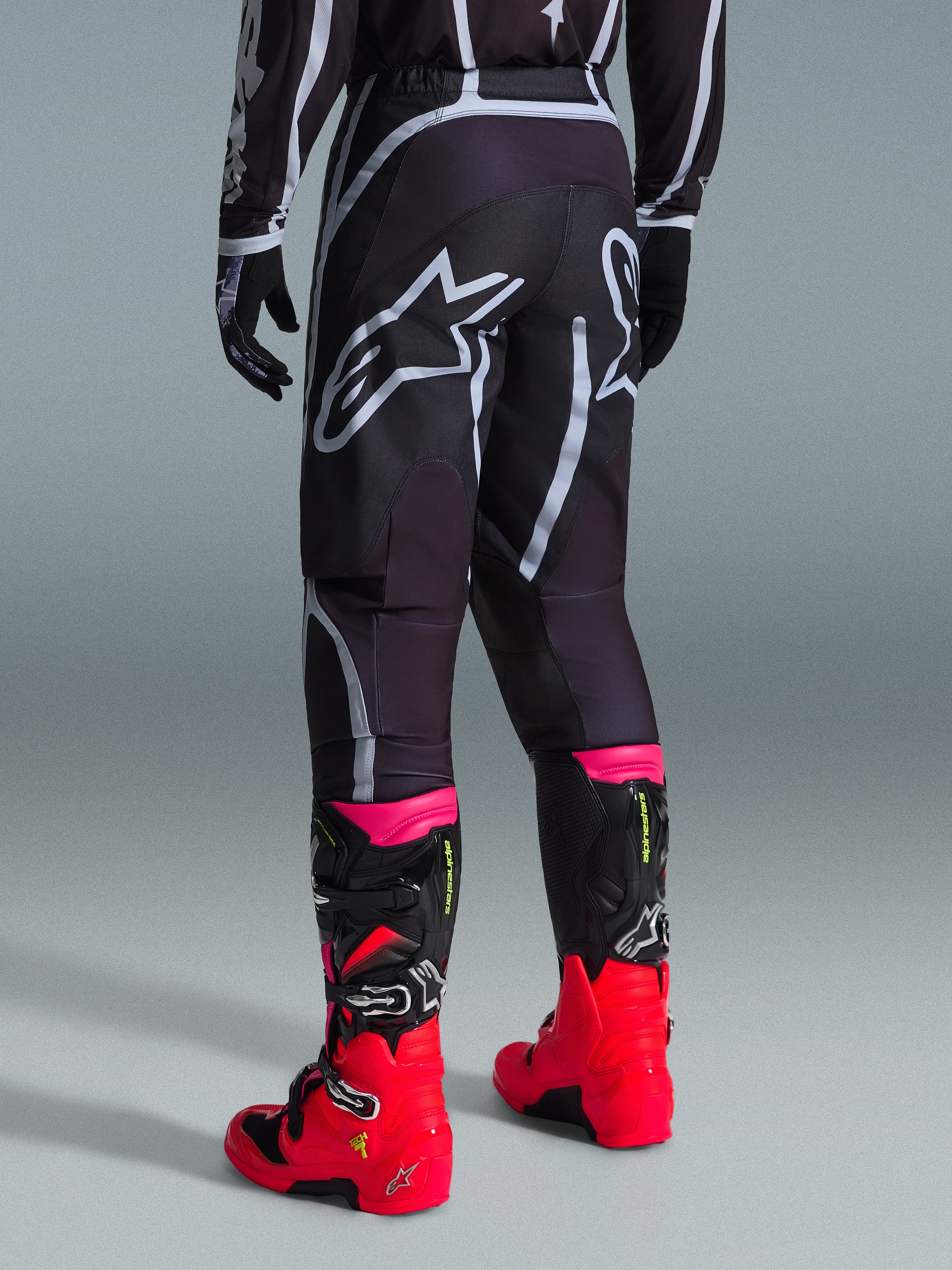 Pantalones Alpinestars Fluid Apex 2026, pantalones de motocross, negro y gris, vista trasera con grandes logotipos blancos de Alpinestars en el asiento, usados con botas en rojo brillante y rosa, construcción duradera de polifabric 600D para rendimiento off-road