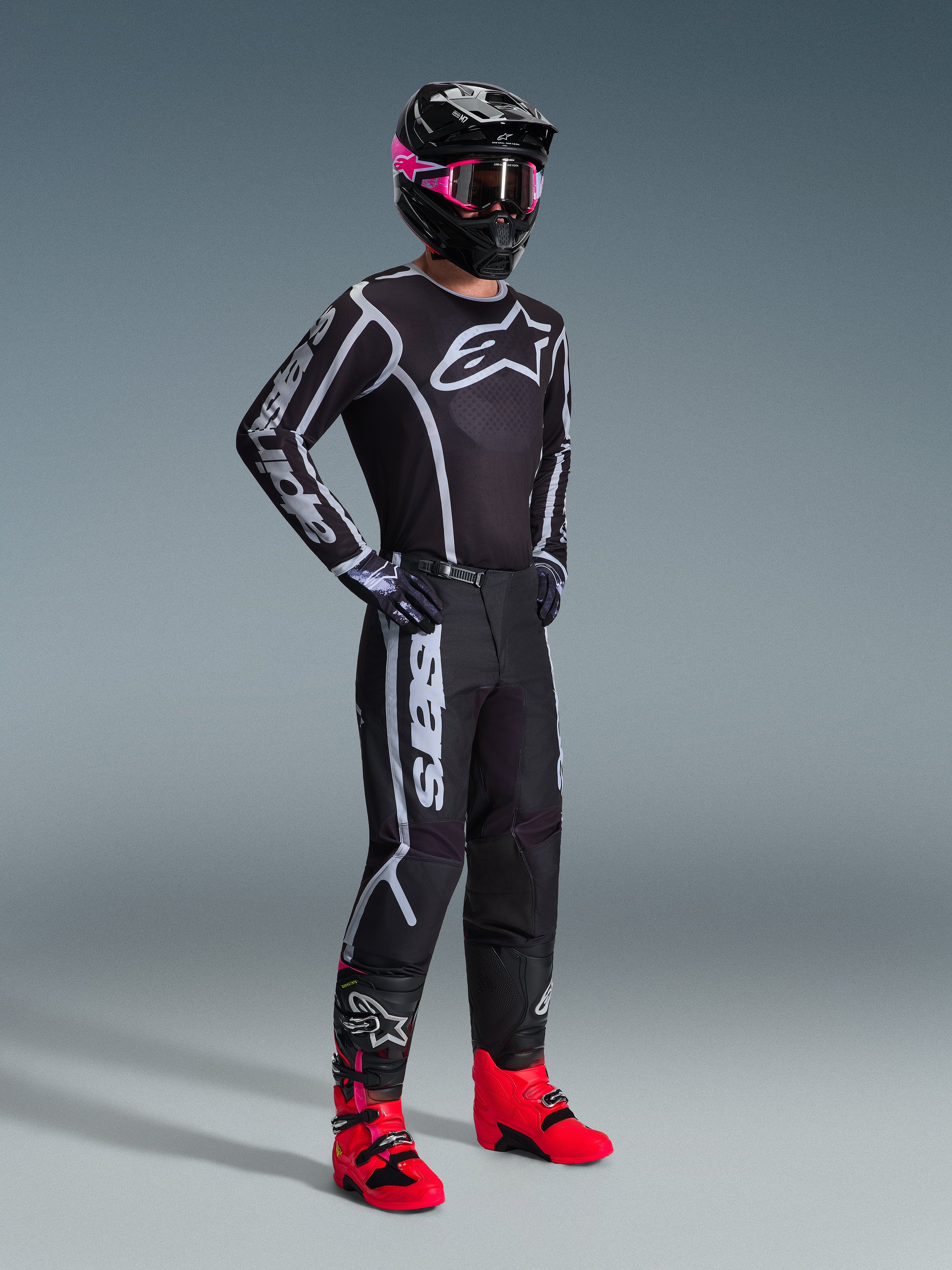 Pantalones Alpinestars 2026 Fluid Apex, pantalones de motocross, negro y gris, combinados con un jersey negro y blanco a juego, casco negro con gafas rosas y botas de color rojo brillante, con logotipos blancos llamativos en las piernas y un diseño de ajuste deportivo para carreras todoterreno