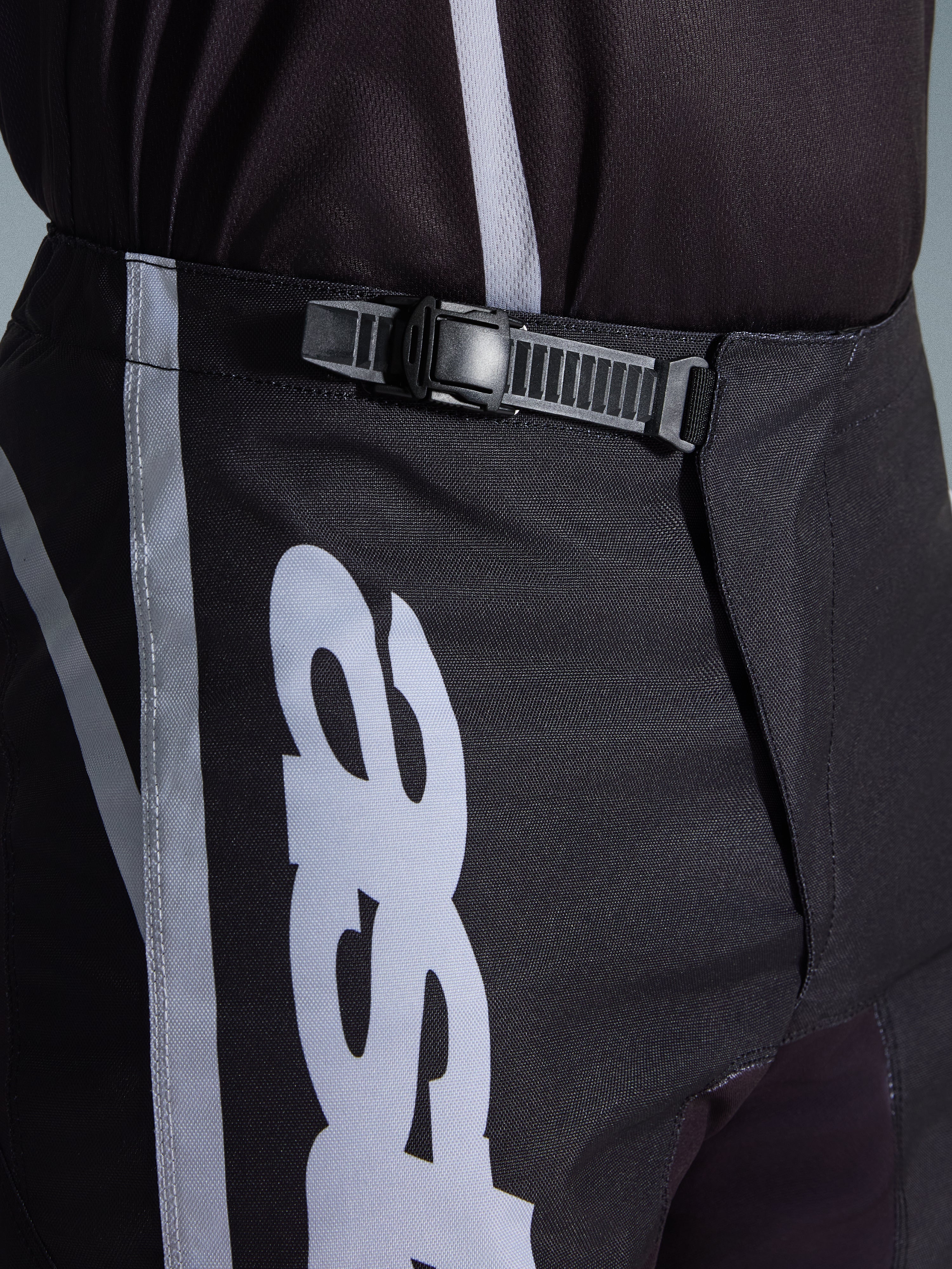 Alpinestars 2026 Fluid Apex Pants, pantalones de motocross, negro y gris, detalle en primer plano del sistema de cierre de trinquete integrado para un ajuste seguro, con gráficos del logotipo en blanco sobre polifabric 600D duradero