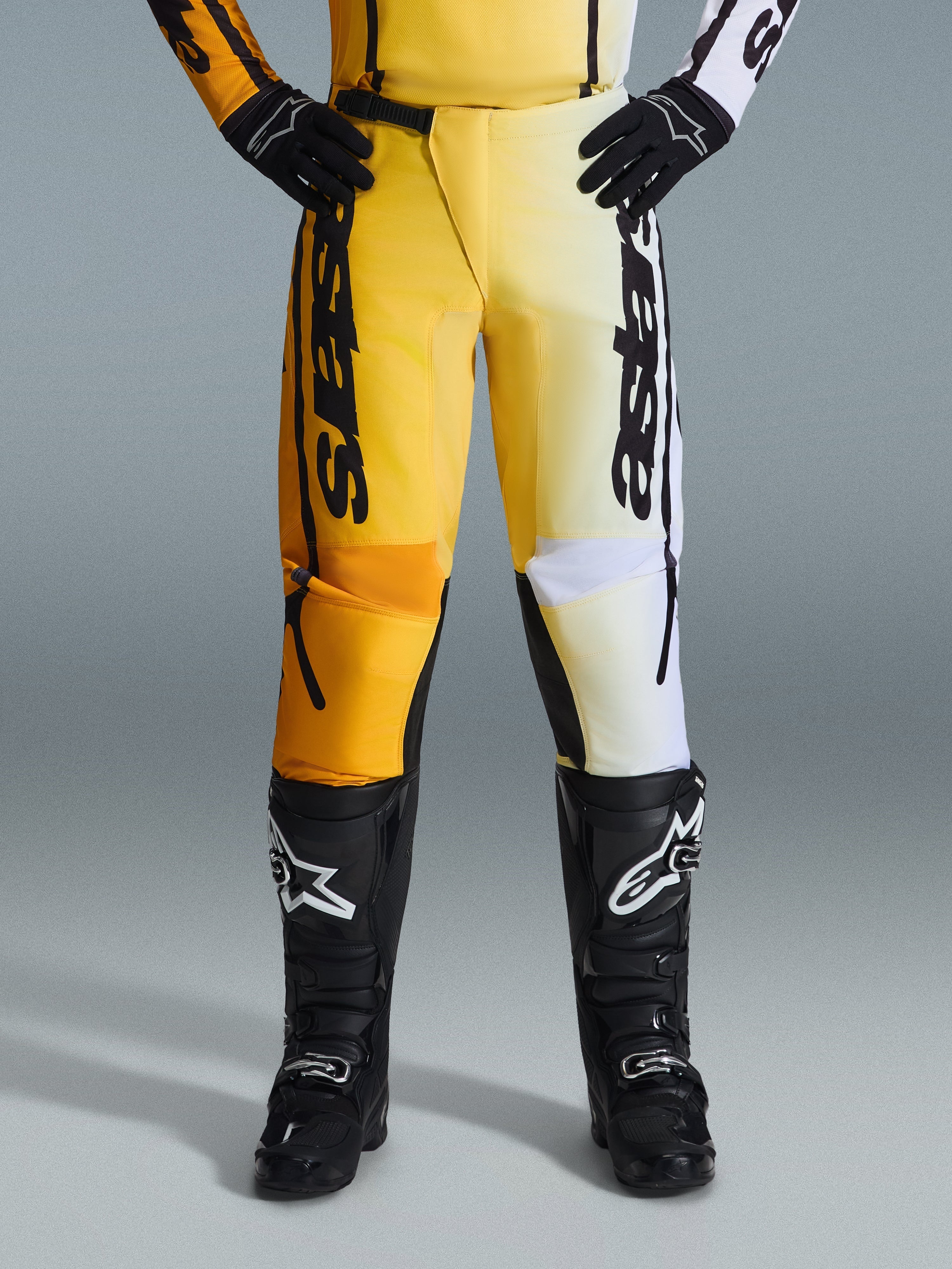 Alpinestars 2026 Fluid Apex Pants, pantalones de motocross, amarillos y negros, diseño de colores divididos con paneles amarillos y blancos, marca vertical 'astars' llamativa, usados con botas y guantes de motocross negros, equipamiento de alto rendimiento para conducción off-road